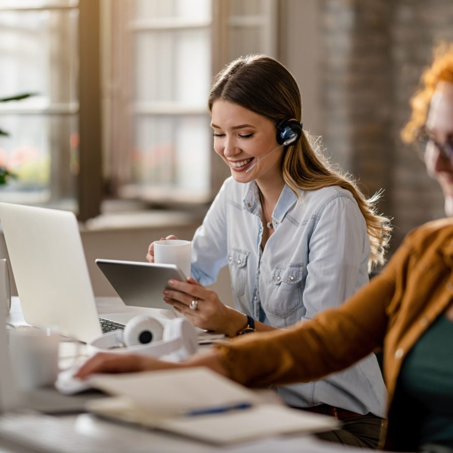 Young happy businesswoman using digital tablet while drinking co Happy female entrepreneur with headset drinking coffee while surfing the net on touchpad in the office.